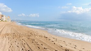Ein sandiger Strand mit Fußspuren, die zum Wasser führen, ein klarer Himmel und Gebäude im Hintergrund.