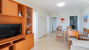Ein Wohnzimmer mit Fernseher, Holzkredenz, Essbereich mit Tisch und Stühlen sowie ein Flur zu einem weiteren Zimmer.