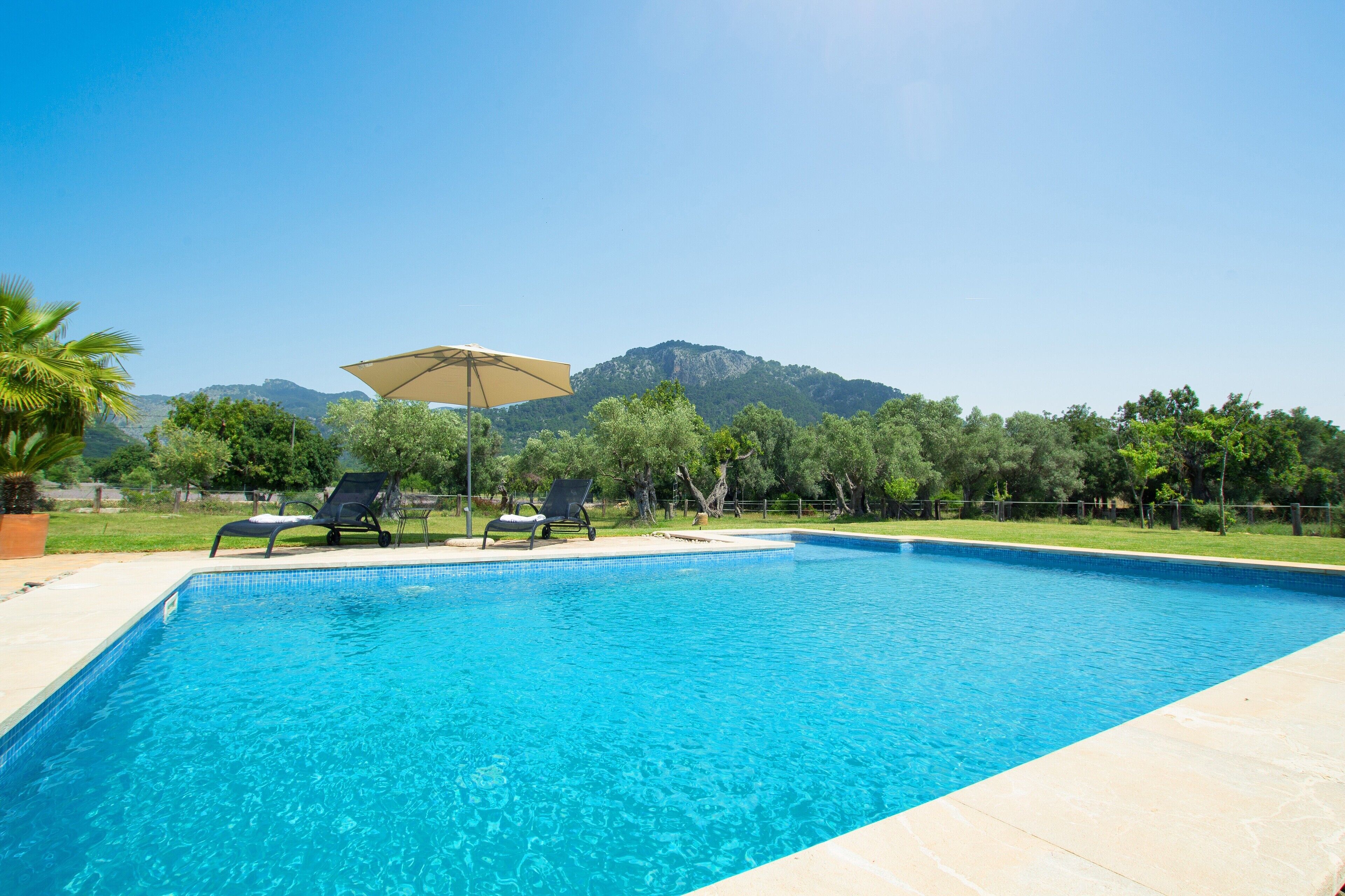 A rectangular swimming pool with clear blue water, surrounded by a tiled deck. There are two lounge chairs and a large umbrella providing shade. The background features a mountainous landscape with trees and a clear sky.