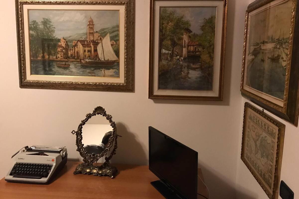 A room with a desk, a typewriter, a mirror, and three framed paintings on the wall.