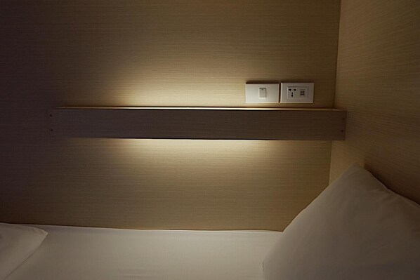 Habitación de hotel con cabecera de madera, mesita de noche con lámpara y toma de corriente.