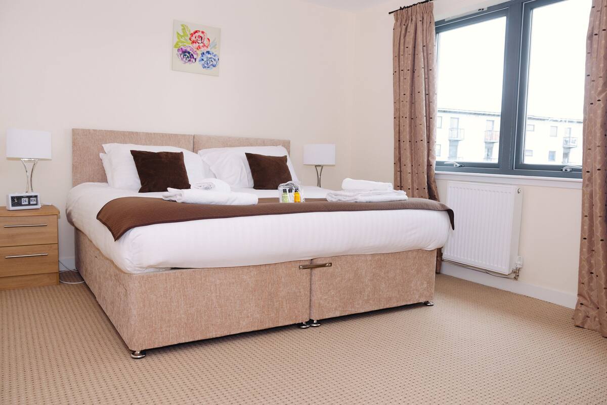 A neatly made bed with a brown headboard, two bedside lamps, a small table with a clock, and a window with curtains.