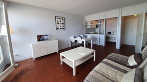 A modern living room with a sofa, coffee table, and dining area. There is a television on a cabinet, a wall-mounted picture frame, and a kitchen area with cabinets and a microwave. - 2-room apartment, sleeps 5 with balcony - Saint-Jean-de-Monts (Saint-Jean-de-Monts)