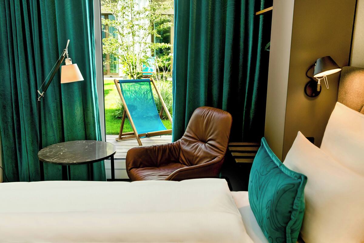 A hotel room with a bed, a brown leather chair, a small table, and a view of a garden through a glass door.