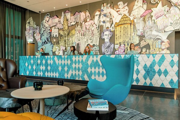 Un vestíbulo con un mural grande de varios paisajes urbanos y figuras, un mostrador de recepción, zona de estar con una mesa redonda y un sillón azul.