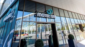 Una entrada de hotel moderna con grandes ventanales de vidrio, un letrero llamativo que dice "Motel One", y un cielo despejado arriba.