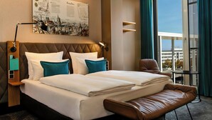 Una habitación de hotel moderna con una cama grande, un banco, un escritorio y vistas a la ciudad.