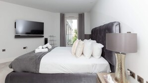A modern hotel room with a large bed, a flat-screen TV mounted on the wall, a bedside table with a lamp, and a view of the outdoors through a window. - Luxurious Central Edinburgh Apartment - Sleeps 6 (Edinburgh)