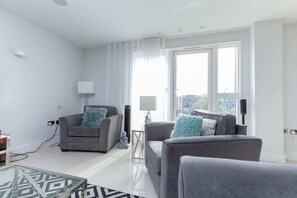 Un salon moderne avec un canapé gris, un fauteuil gris plus petit et une table basse en verre. Une grande fenêtre est agrémentée de rideaux, et on y trouve une lampe ainsi qu’un haut-parleur fixé au mur.