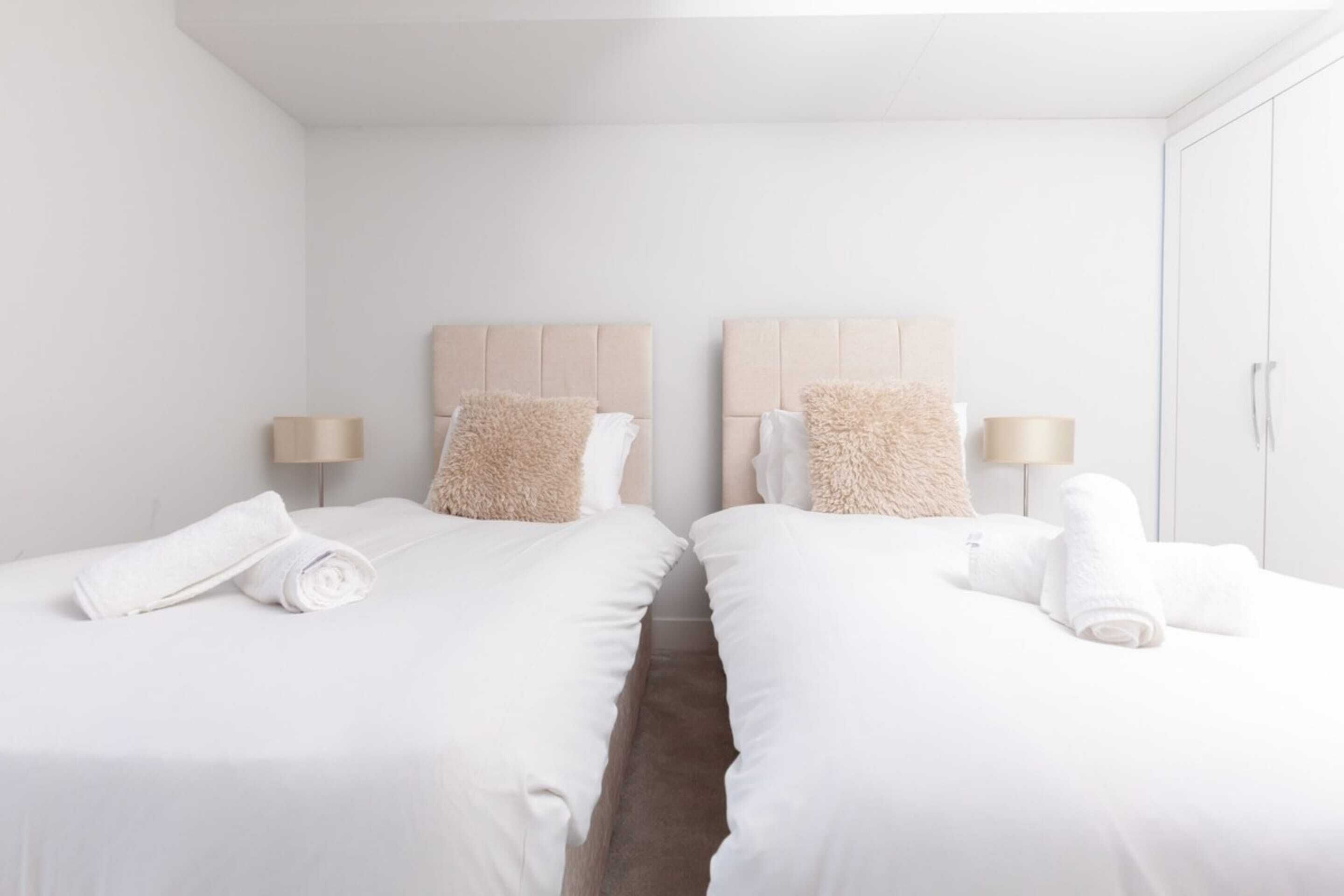 Quarto de hotel com duas camas, cada uma com cabeceira, roupa de cama branca e uma toalha enrolada. Há uma mesa de cabeceira com abajur e um guarda-roupa com espelho.