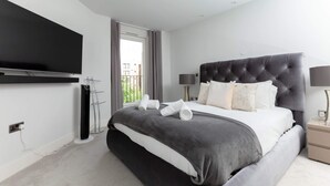 A modern hotel room with a large bed, a flat-screen TV mounted on the wall, a bedside table with a lamp, and a view of the outdoors through a window. - Luxurious Central Edinburgh Apartment - Sleeps 6 (Edinburgh)