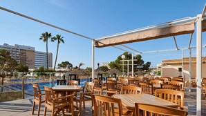 A rooftop pool area with wooden tables and chairs, a covered seating area, and a view of the surrounding buildings and palm trees. -    AluaSun Cala Antena – All Inclusive (Manacor)