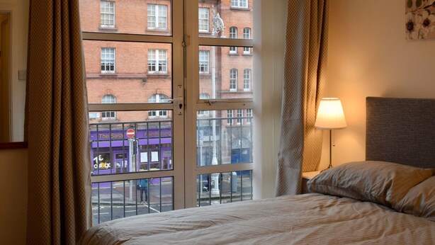 A hotel room with a bed, a lamp, and a view of a brick building with a salon.