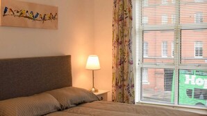 A bedroom with a bed, a nightstand, a lamp, a floral curtain, and a window with a view of buildings.