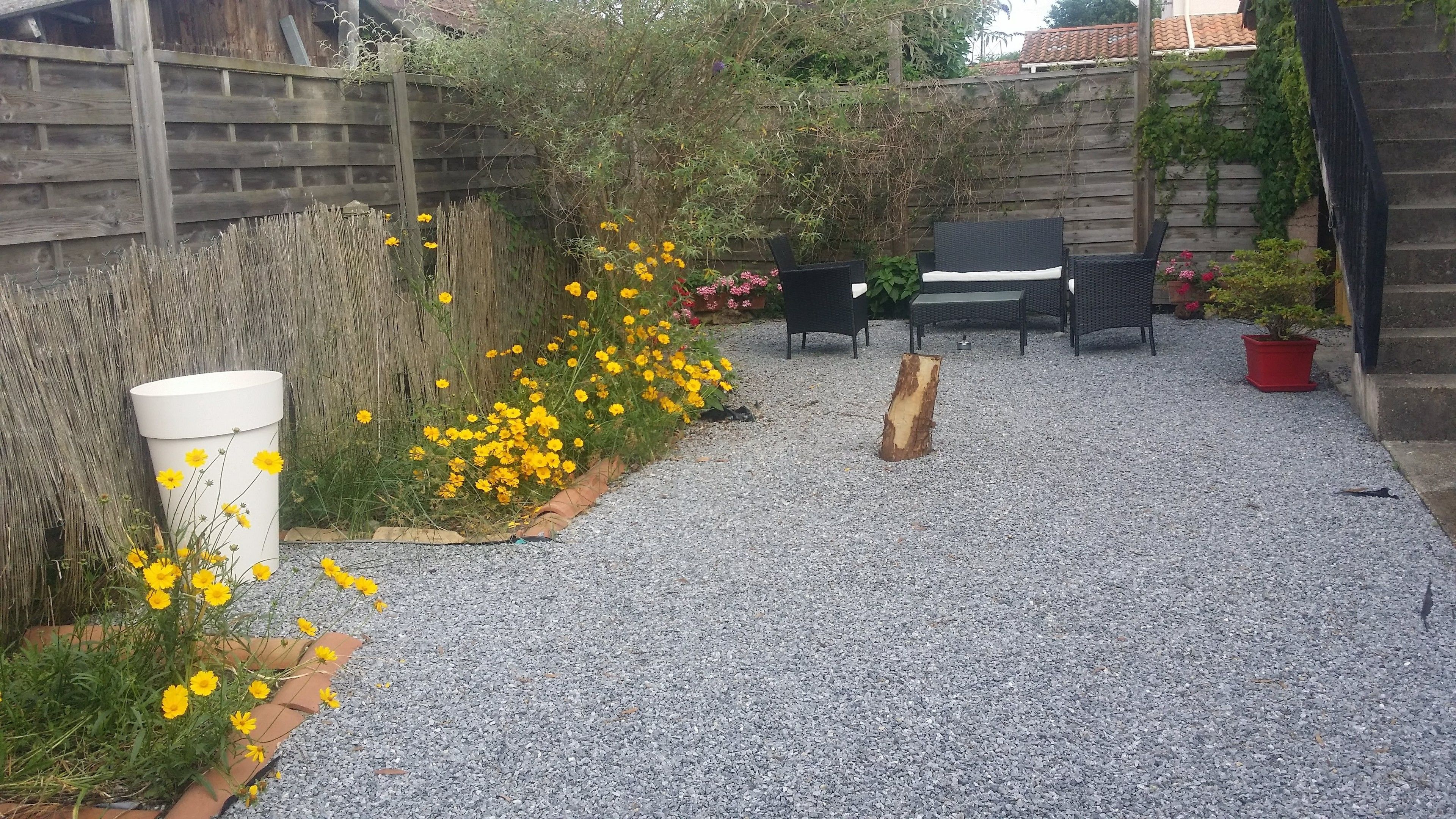 Eine Pflasterterrasse mit Holzlattenzaun, ein weißer Pflanzkasten mit gelben Blumen und Gartenmöbeln.