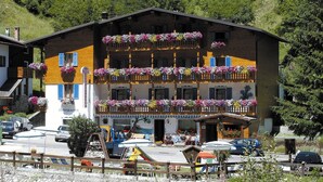 A multi-story hotel with balconies adorned with colorful flowers, a visible entrance, and a parking area in front. - Albergo Genzianella (Rocca Pietore)