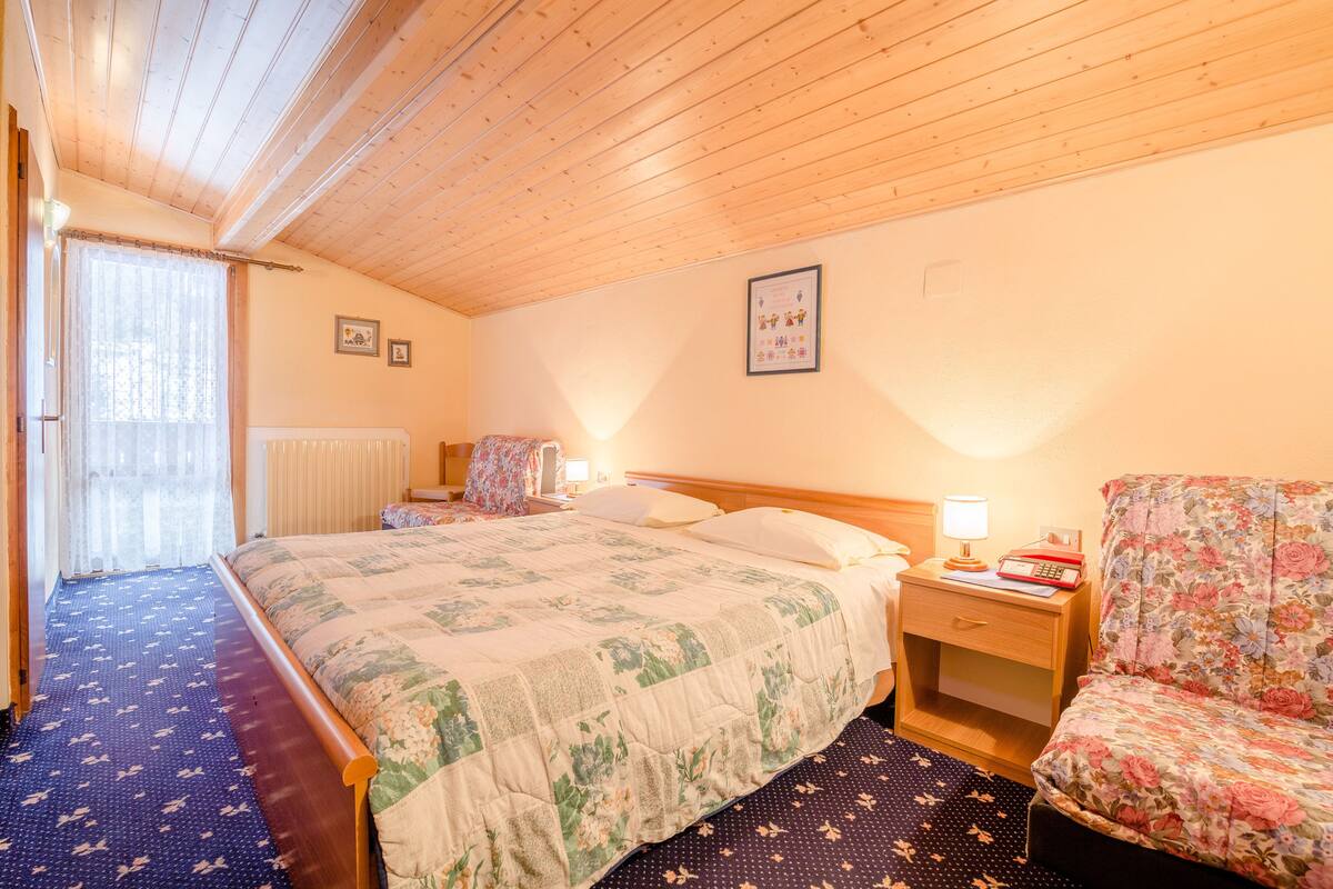 A bedroom with a wooden ceiling, a bed with floral bedding, a nightstand with a lamp, and a chair.