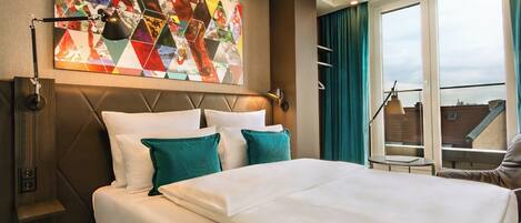 A hotel room with a large bed, a headboard with geometric patterns, a colorful abstract artwork on the wall, and a view of the city through the window.