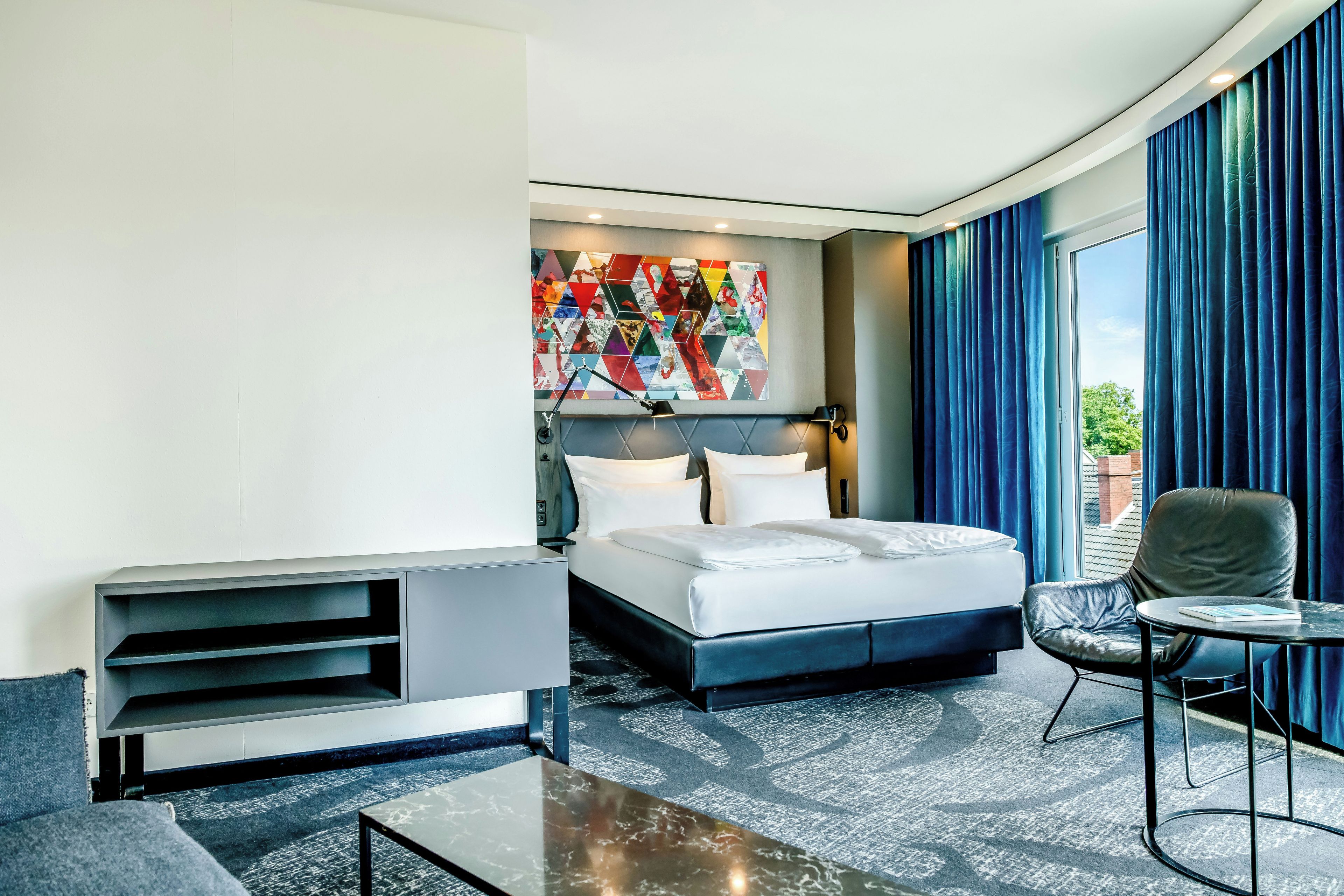 A hotel room with a bed, a seating area, a wall art piece, and a view through the window.