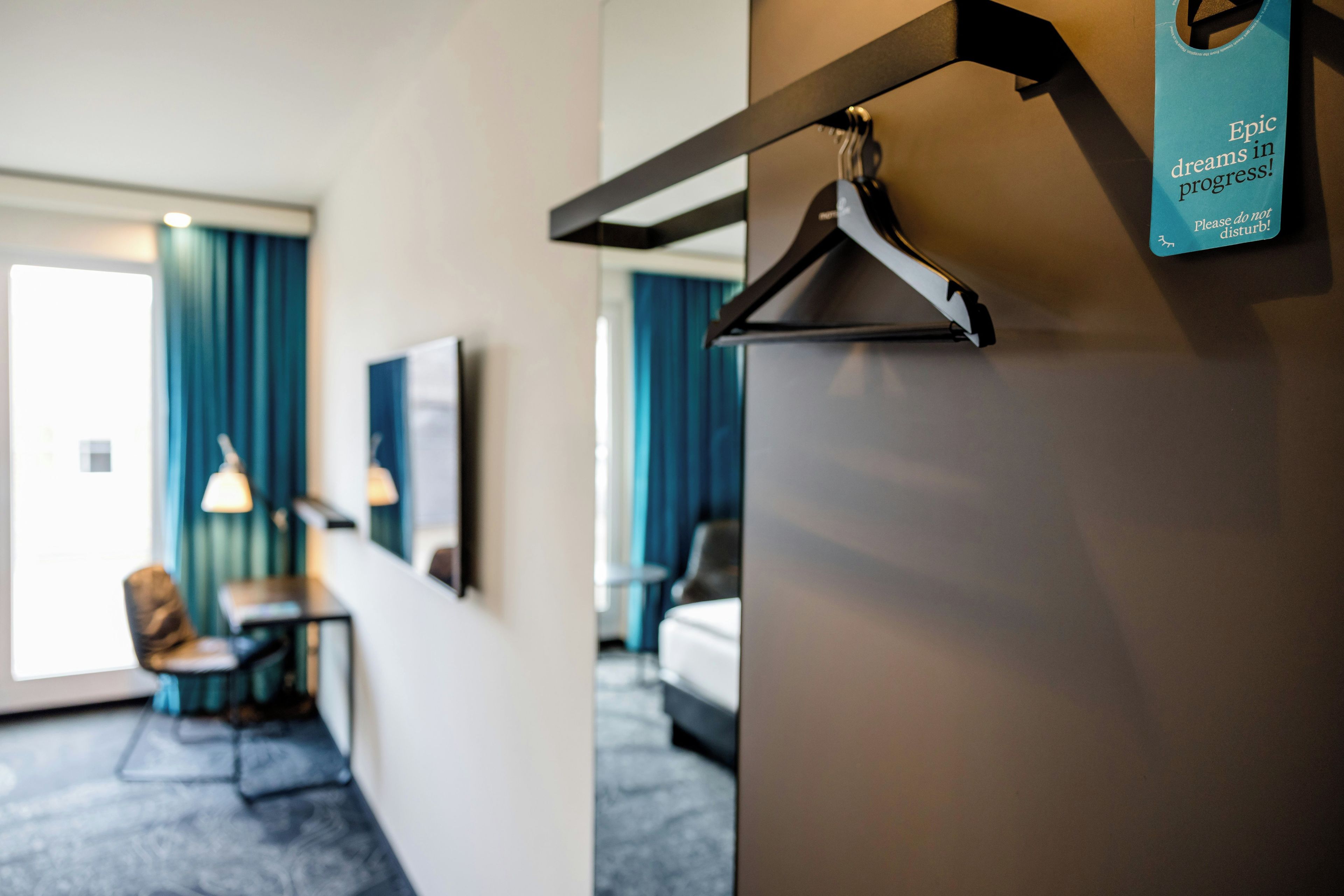 A hotel room with a bed, a desk, and a chair. There is a coat hanger on the wall.
