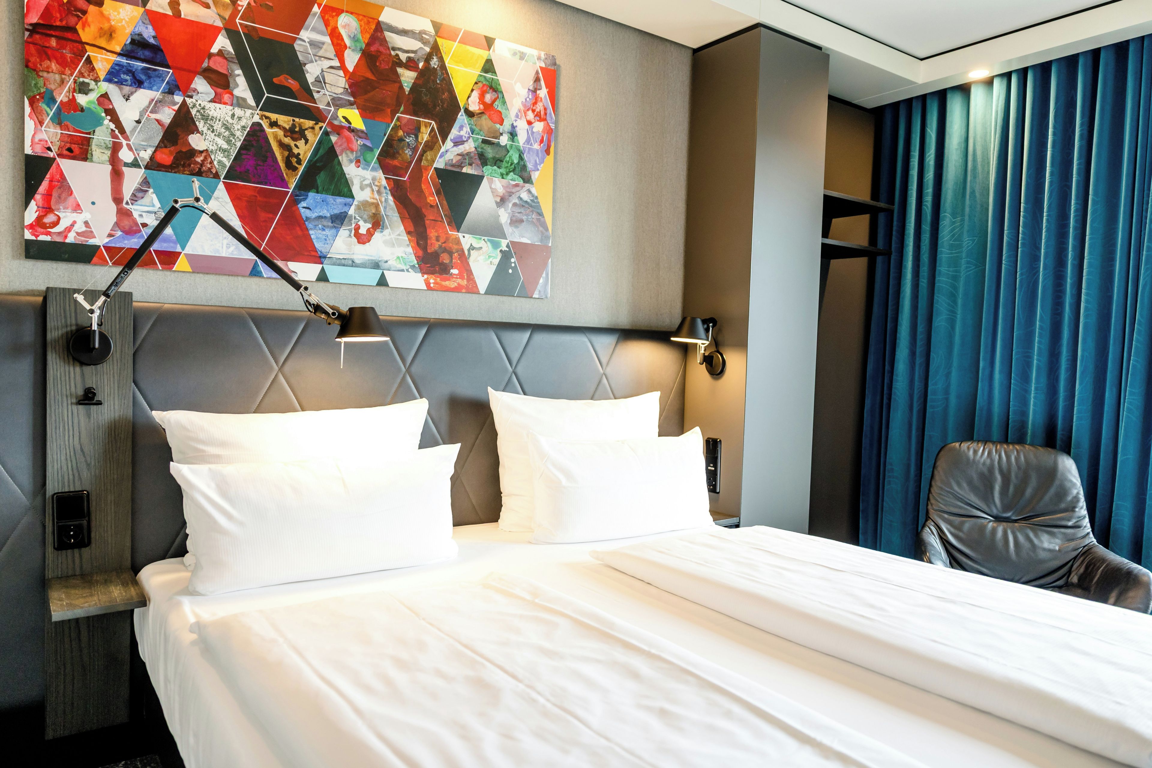 A hotel room with a bed, pillows, a headboard, a wall art piece, a desk lamp, a chair, and a window with curtains.