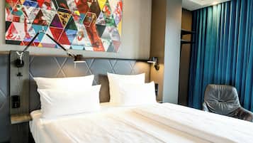 A hotel room with a bed, pillows, a headboard, a wall art piece, a desk lamp, a chair, and a window with curtains.