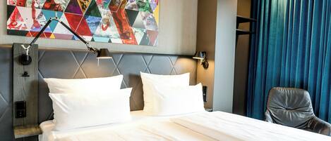 A hotel room with a bed, pillows, a headboard, a wall art piece, a desk lamp, a chair, and a window with curtains.