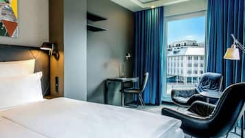 Ein Hotelzimmer mit Bett, Schreibtisch, Stuhl und Blick auf die Stadt durch große Fenster.
