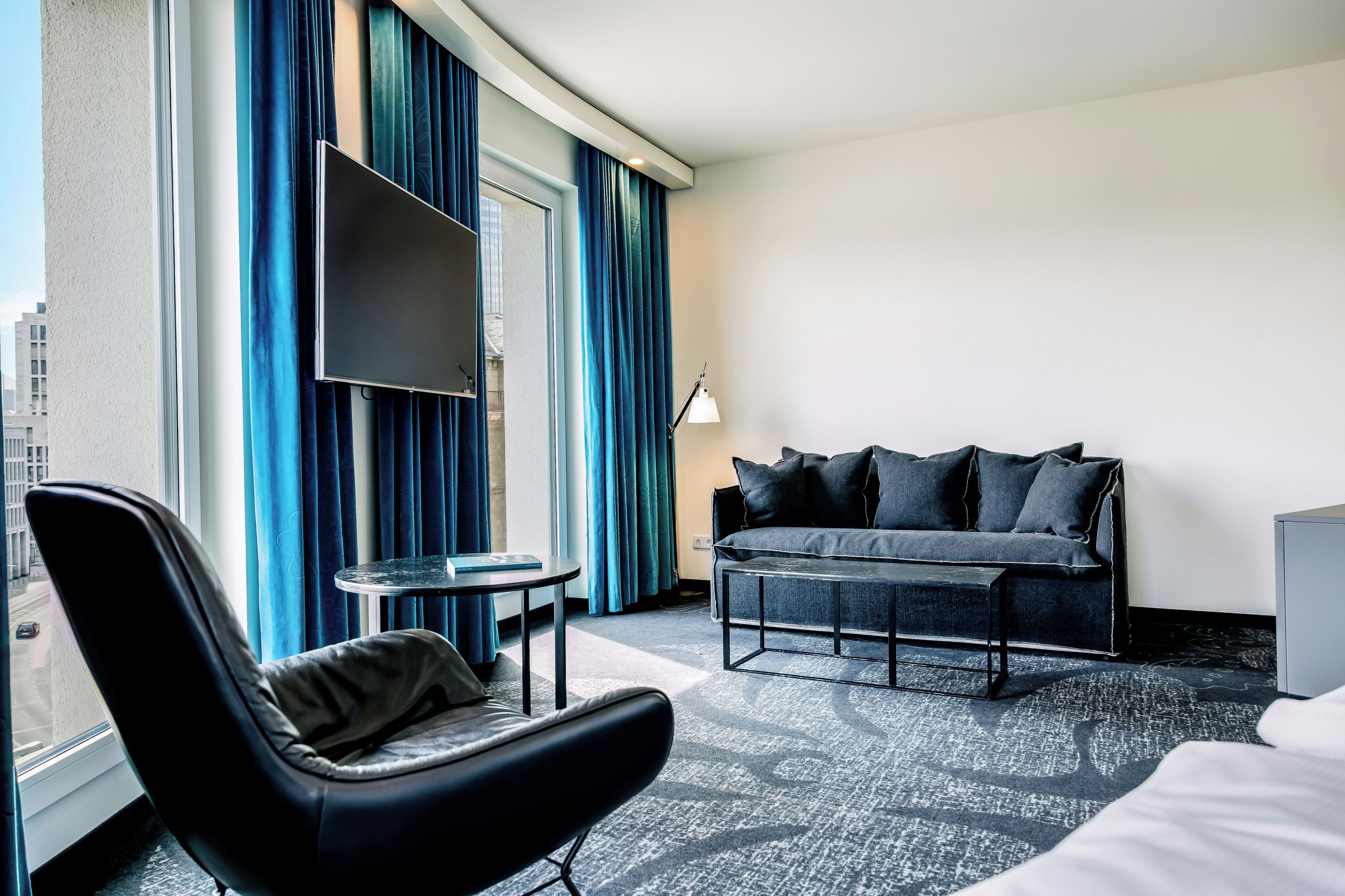 A hotel room with a black leather chair, a sofa with multiple cushions, a small round table, and a flat-screen TV mounted on the wall.