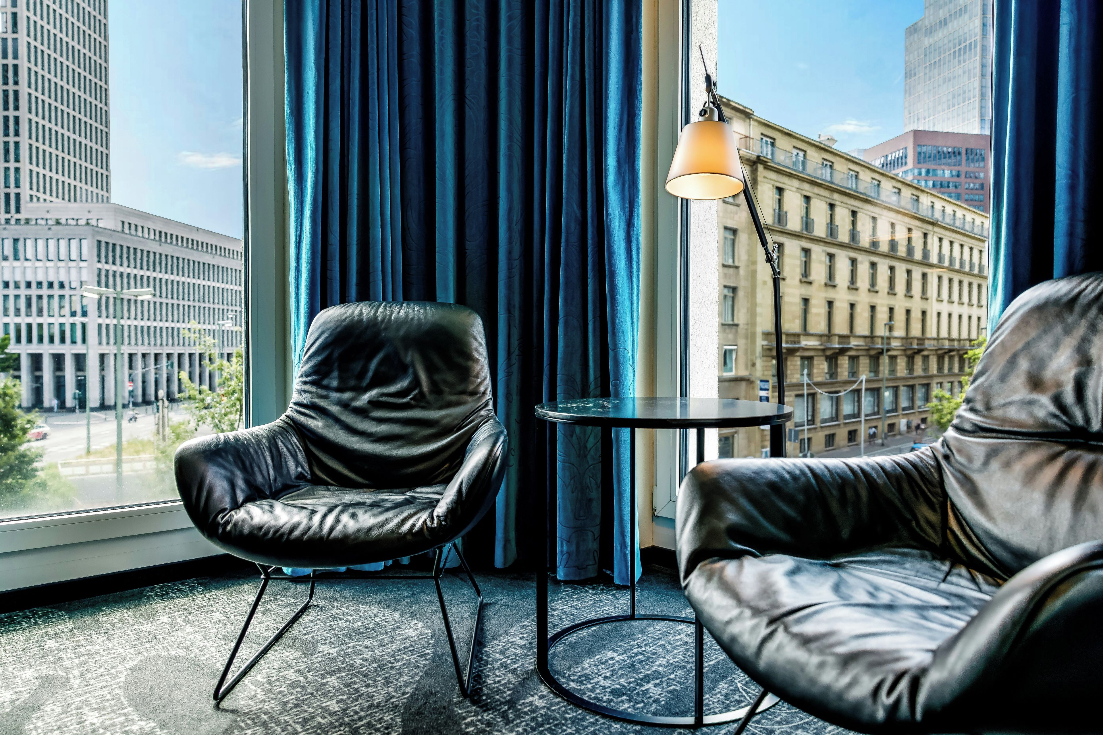 Habitación con dos sillones de cuero, una mesita y vistas a edificios de la ciudad a través de amplias ventanas.
