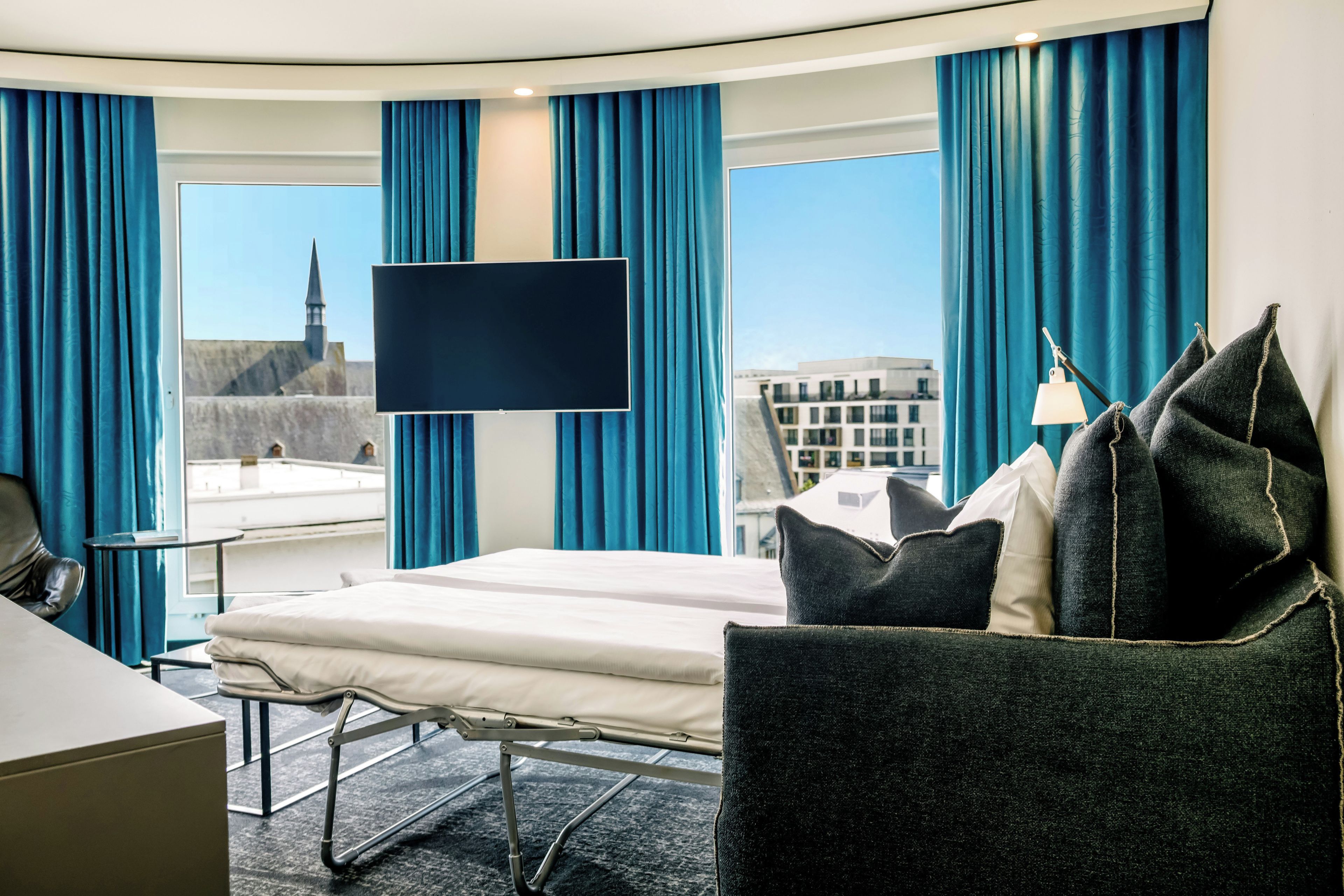 Ein Hotelzimmer mit einem Bett, einem Schreibtisch, einem Stuhl, einem Fernseher und Blick auf Gebäude sowie einen Kirchturm.