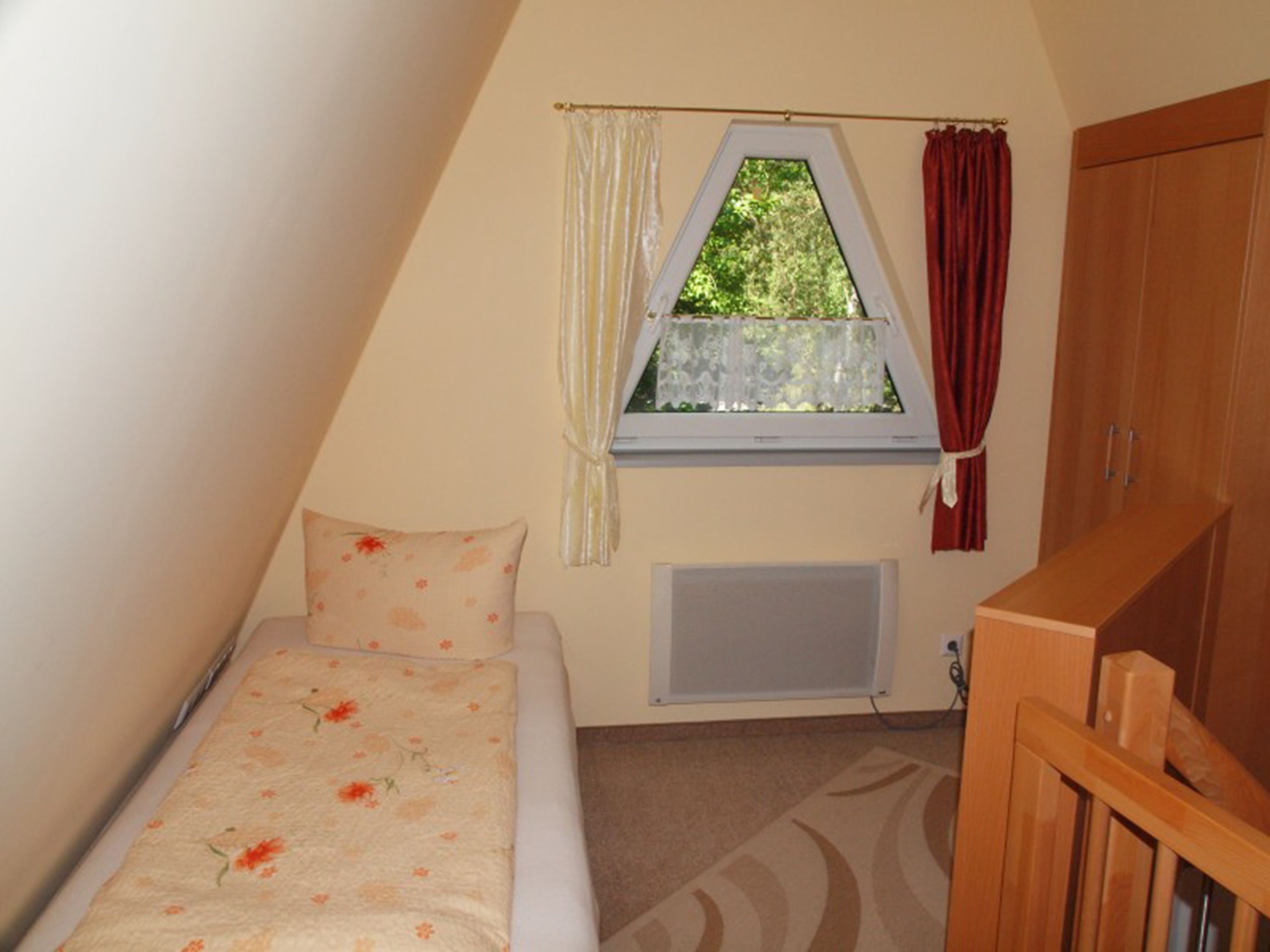 A small, triangular bedroom with a single bed, a window with curtains, a radiator, and a wooden wardrobe.