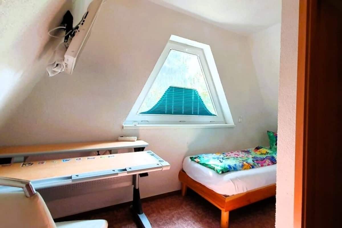 Une petite chambre mansardée de forme triangulaire, avec un plafond en pente, un lit simple, un bureau et une fenêtre donnant sur le ciel.