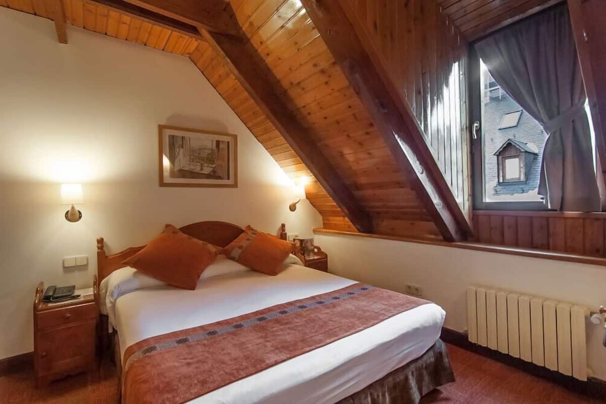 Ein Schlafzimmer mit geneigter Decke, Holzbalken, einem Bett mit weißer Tagesdecke und orangefarbenen Kissen, einem Nachttisch mit Telefon, einem gerahmten Bild an der Wand und einem Fenster mit Vorhängen.