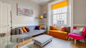 Ein Wohnzimmer mit grauem Sofa, orangefarbenem Hocker und einem rosafarbenen Sessel. Es gibt einen Glastisch, eine Lampe und Kunstwerke an den Wänden.