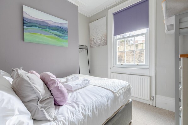 A bedroom with a bed, pillows, a window with blinds, a radiator, and a wall with a painting.