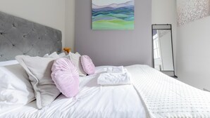 Ein ordentlich bezogenes Bett mit weißen Leinen und rosafarbenen Kissen, ein grauer Stoffkopfteil und ein Gemälde an der Wand.