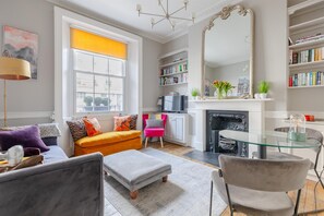 Un salon moderne avec un canapé, un pouf et une table en verre. On y trouve également une cheminée, une bibliothèque et un grand miroir.