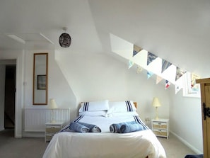 A bedroom with a bed, two bedside tables, a radiator, a hanging light fixture, a window with a view, and a door.