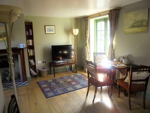 A living room with a television on a stand, a bookshelf, a dining table with chairs, and a window with curtains.