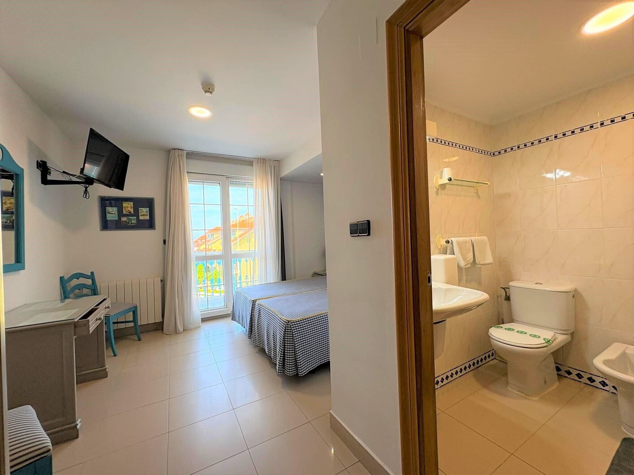 Habitación de hotel con cama, escritorio, silla, televisor montado en la pared, baño con inodoro y lavabo, y ventana con cortinas.