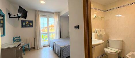 Habitación de hotel con cama, escritorio, silla, televisor montado en la pared, baño con inodoro y lavabo, y ventana con cortinas.