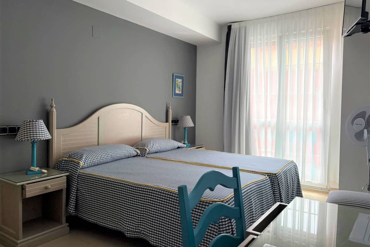 A bedroom with a bed, two bedside tables, a nightstand, a window with curtains, and a wall-mounted fan.