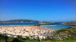 Una playa concurrida, con aguas azul turquesa, afloramientos rocosos y una colina distante con edificios.