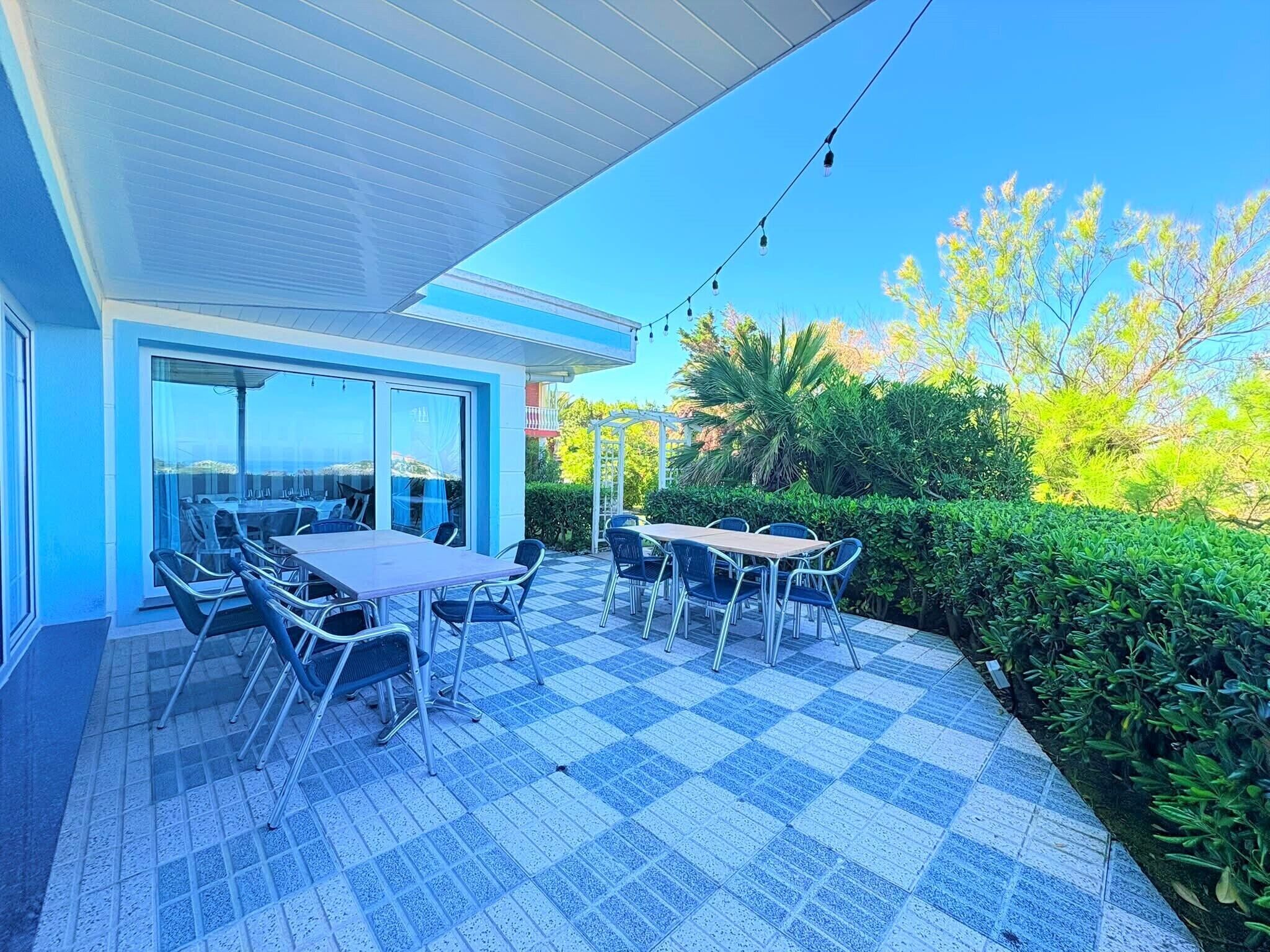 Eine überdachte Terrasse mit Sitzgelegenheiten und Meerblick, umgeben von einer Hecke.