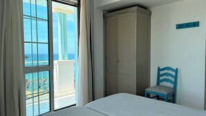 Habitación de hotel con cama, armario, una silla y vista al océano a través de amplios ventanales.