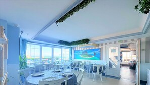 Un comedor con mesas redondas, sillas y un amplio ventanal con vistas al mar.