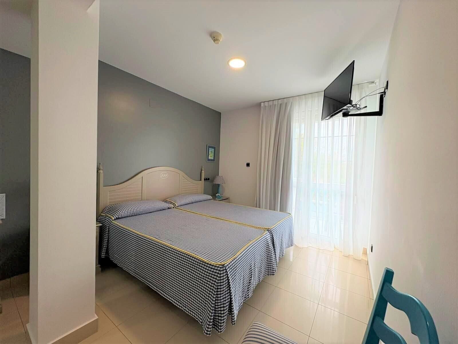 Een hotelkamer met twee bedden, een aan de muur bevestigde televisie, een raam met gordijnen en een betegelde vloer.
