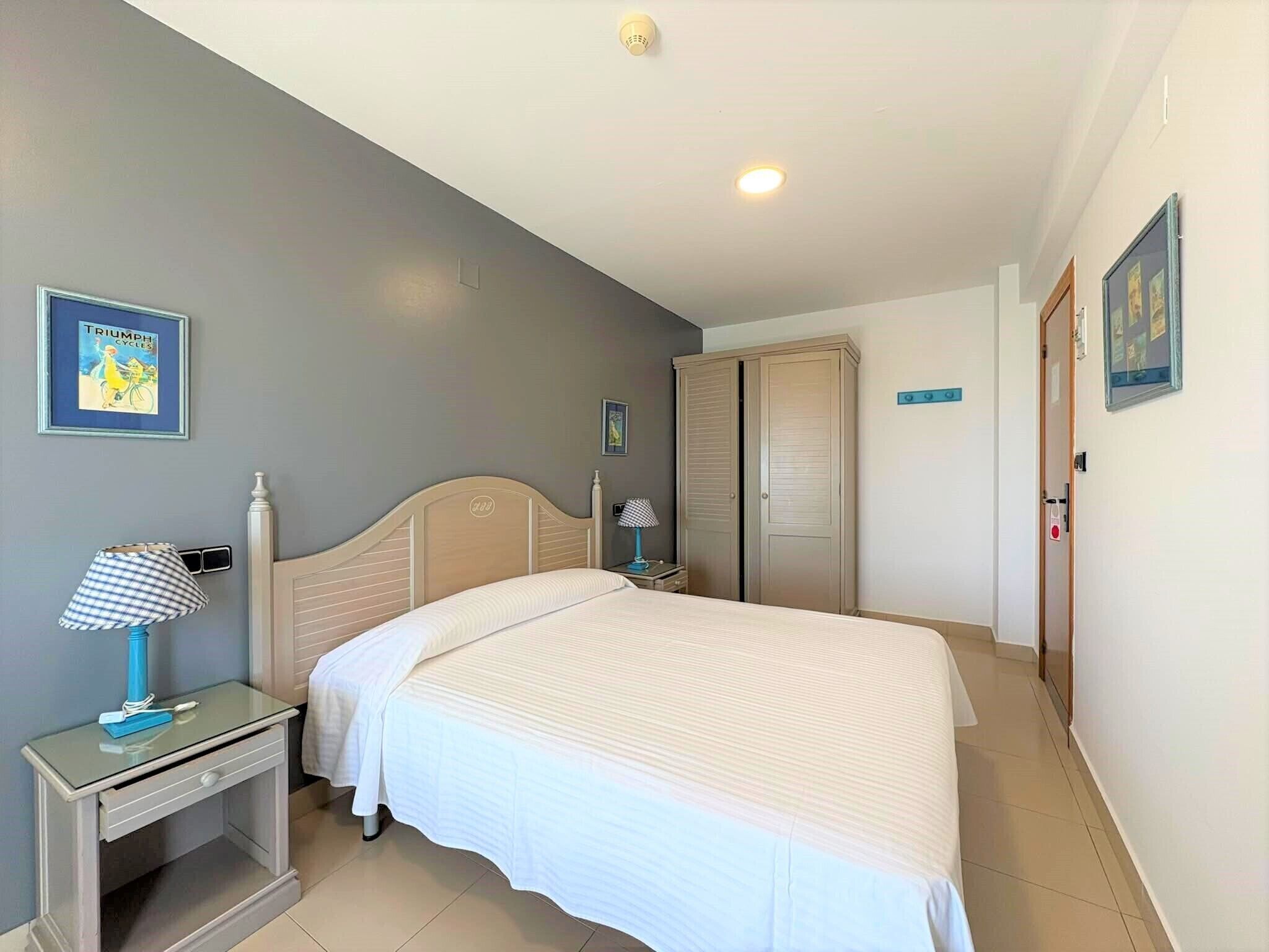Un dormitorio con cama, mesita de noche, lámpara, armario y un cuadro en la pared.