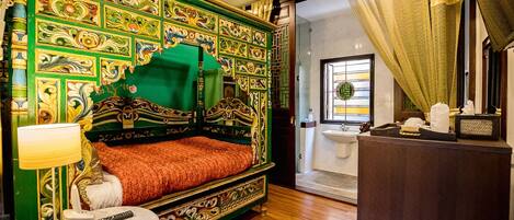 A room with a green and gold ornate bed, a round table with a lamp, and a view of a bathroom through an open door.